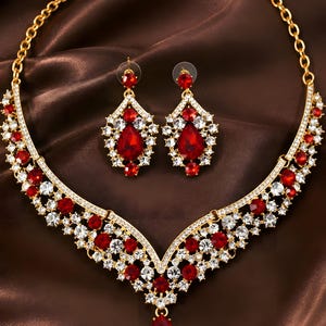 May include: A gold-tone necklace and earring set featuring teardrop-shaped red crystals and clear round rhinestones. The necklace has a V-shaped centerpiece with a dangling red crystal, while the earrings mirror the design with matching red and clear stones.