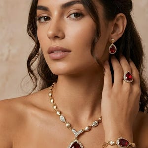 May include: A jewelry set featuring a gold-tone necklace, bracelet, ring, and drop earrings, each centered with a large, pear-shaped deep red gemstone surrounded by small clear crystals. The necklace chain consists of alternating gold-tone links.