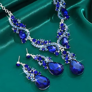 Può includere: Un set di gioielli color argento composto da una collana e orecchini pendenti abbinati. Ogni pezzo presenta grandi cristalli blu scuro a forma di goccia circondati da cristalli rotondi blu chiaro incastonati in una montatura in metallo.