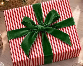 May include: A square gift box wrapped in red and white striped paper, tied with a wide, dark green velvet ribbon bow. The gift is positioned near a Christmas tree with ornaments, a white candle in a glass holder, and snowflake cookies.