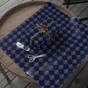 May include: A rectangular gift box wrapped in dark blue paper with a diamond argyle pattern featuring small nutcracker illustrations, tied with a tan ribbon bow, accompanied by a clear tape dispenser and silver scissors.