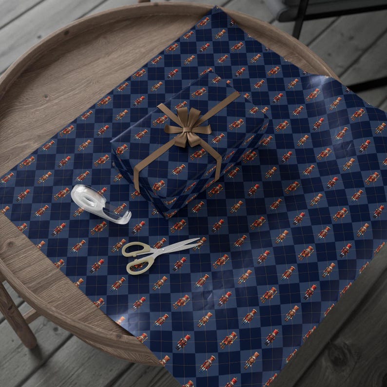 May include: A rectangular gift box wrapped in dark blue paper with a diamond argyle pattern featuring small nutcracker illustrations, tied with a tan ribbon bow, accompanied by a clear tape dispenser and silver scissors.