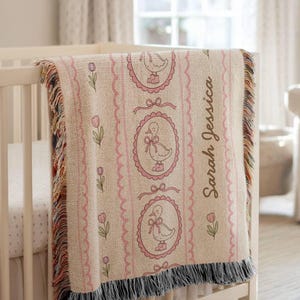May include: A beige baby blanket with a pink duck design and the name "Sarah Jessica" printed on it. The blanket has a fringed edge and is draped over a white crib. Pink tulips and bows decorate the blanket.