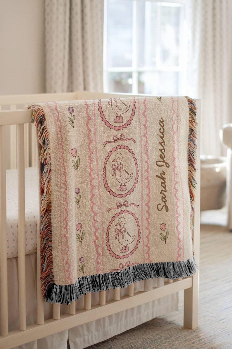 May include: A beige baby blanket with a pink duck design and the name "Sarah Jessica" printed on it. The blanket has a fringed edge and is draped over a white crib. Pink tulips and bows decorate the blanket.