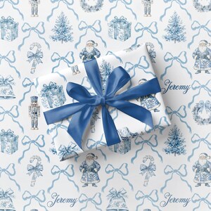May include: A wrapped present with a large blue satin bow, set against a white background with a repeating pattern of blue Christmas illustrations. The pattern includes Santa Claus figures, gift boxes, Christmas trees, and bows. The name "Jeremy" is printed on the wrapping paper.
