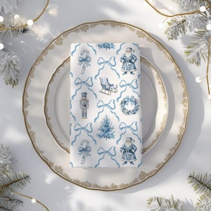 May include: A folded white napkin with a blue Christmas pattern, including Santa Claus, bows, Christmas trees, and nutcrackers. The napkin sits on a white plate with a gold rim, creating a festive holiday table setting.