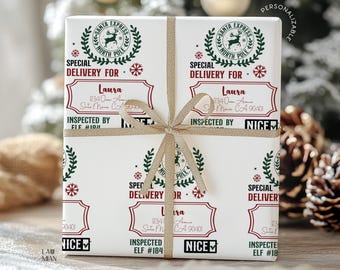 May include: A white gift box wrapped with a gold ribbon, featuring a repeating Santa Express North Pole print in red and green. The design includes labels with the text Special Delivery For, Laura, 1234 Ocean Avenue, Santa Monica, CA 90401, and Inspected By Elf #184 Nice.