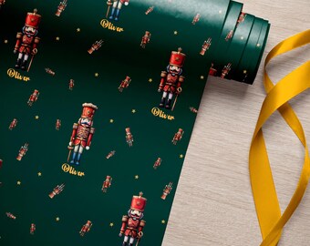 May include: A roll of dark green wrapping paper featuring a repeating pattern of red and gold nutcracker figures and small yellow stars. The name Oliver is printed in gold script beneath each nutcracker. A yellow ribbon lies to the side.