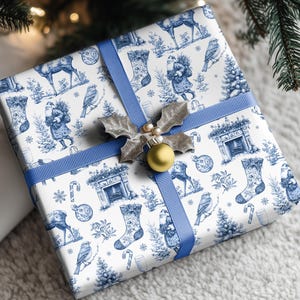 Puede incluir: Una caja de regalo cuadrada envuelta en papel blanco con un estampado azul estilo toile que muestra renos, calcetines, p&aacute;jaros y chimeneas. La caja est&aacute; atada con una cinta azul y decorada con un adorno de hojas de acebo plateadas.