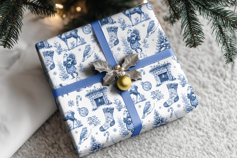 Puede incluir: Una caja de regalo cuadrada envuelta en papel blanco con un estampado azul estilo toile que muestra renos, calcetines, p&aacute;jaros y chimeneas. La caja est&aacute; atada con una cinta azul y decorada con un adorno de hojas de acebo plateadas.
