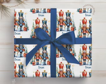 May include: A square gift box wrapped in white paper featuring a repeating pattern of red and blue nutcracker figures, Christmas trees, and gift boxes, with the name Brian printed in blue, tied with a wide navy blue satin ribbon.