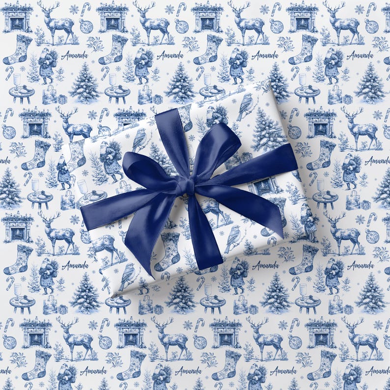 Puede incluir: Una caja de regalo rectangular envuelta en papel blanco con un patr&oacute;n azul repetitivo de estilo toile que muestra ciervos, &aacute;rboles de Navidad, calcetines, chimeneas y el nombre Amanda, atada con un lazo grande de sat&eacute;n azul oscuro.