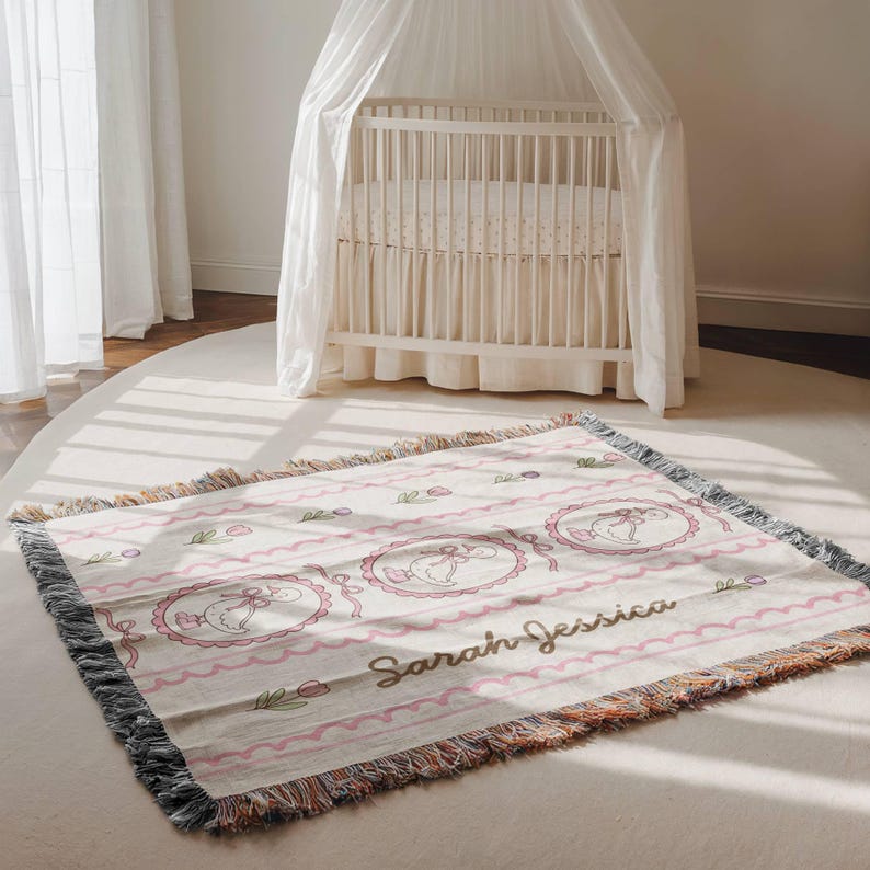 May include: A woven baby blanket with a pink and white design. The blanket features a repeating pattern of a baby in a pink oval frame, pink ribbons, and the name "Sarah Jessica" in cursive. The blanket has a fringed edge.