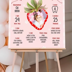 May include: A pink and white sign on an easel, featuring the text "Olivia's berry first year." The sign includes a photo of a baby, measurements in inches and pounds, and favorite things. Strawberries decorate the top.