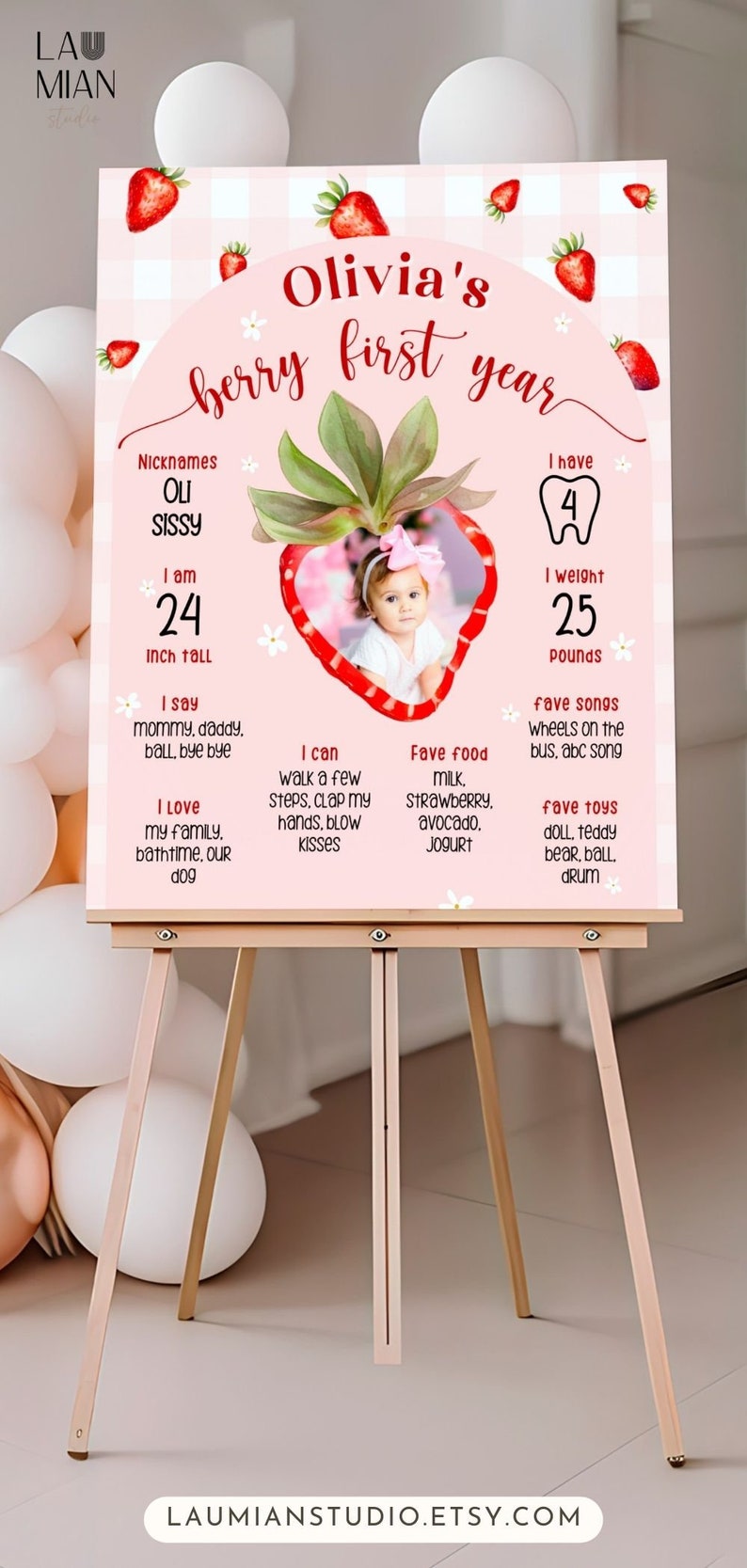 May include: A pink and white sign on an easel, featuring the text "Olivia's berry first year." The sign includes a photo of a baby, measurements in inches and pounds, and favorite things. Strawberries decorate the top.