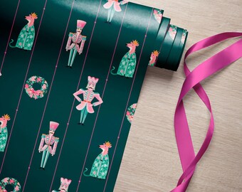 May include: A roll of dark green wrapping paper featuring a repeating pattern of pink nutcracker figures, pink-crowned animals in green robes, and floral wreaths, accented by thin vertical pink lines and a bright pink ribbon.
