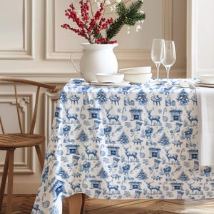 Puede incluir: Un mantel blanco con un estampado azul navideño con renos, árboles de Navidad y calcetines. Un jarrón blanco con bayas rojas y vegetación se encuentra sobre la mesa, junto con cuencos blancos y copas de vino. Se ve una silla de madera.