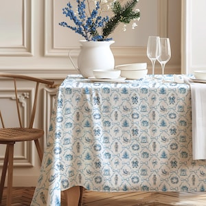 May include: A festive tablecloth with a repeating pattern of blue Christmas-themed illustrations on a white background. The design includes images of Santa, nutcrackers, bows, and Christmas trees. A white vase with blue berries and greenery sits on the table, along with white bowls and wine glasses.