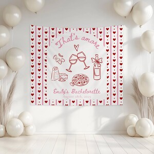 May include: A white backdrop with a red and pink banner that says "That's amore" with a heart and a wine glass clinking with another wine glass. There is a bottle of wine, a pizza, a grater, a piece of cheese, and a bow. The banner also says "Emily's Bachelorette August 18th, 2025".