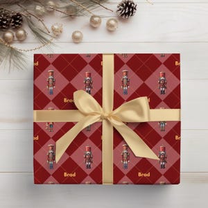 May include: A square gift box wrapped in red and dark red plaid paper featuring a repeating nutcracker pattern and the name Brad printed in gold. The box is tied with a wide, smooth, gold satin ribbon arranged in a cross pattern.