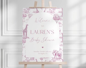 May include: A baby shower sign on a wooden easel. The sign has a white background with a pink toile pattern of giraffes, elephants, and balloons. The text reads "Welcome to LAUREN'S Baby Shower" with the date and location.