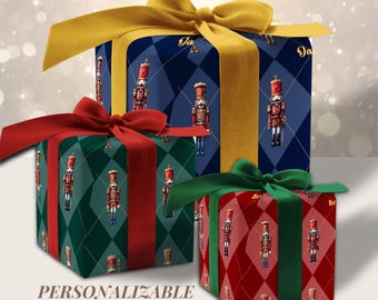 May include: Three square gift boxes wrapped in paper featuring a nutcracker pattern on argyle backgrounds. The largest box is dark blue with a gold ribbon, the green box has a red ribbon, and the red box has a green ribbon. Text reads PERSONALIZABLE.