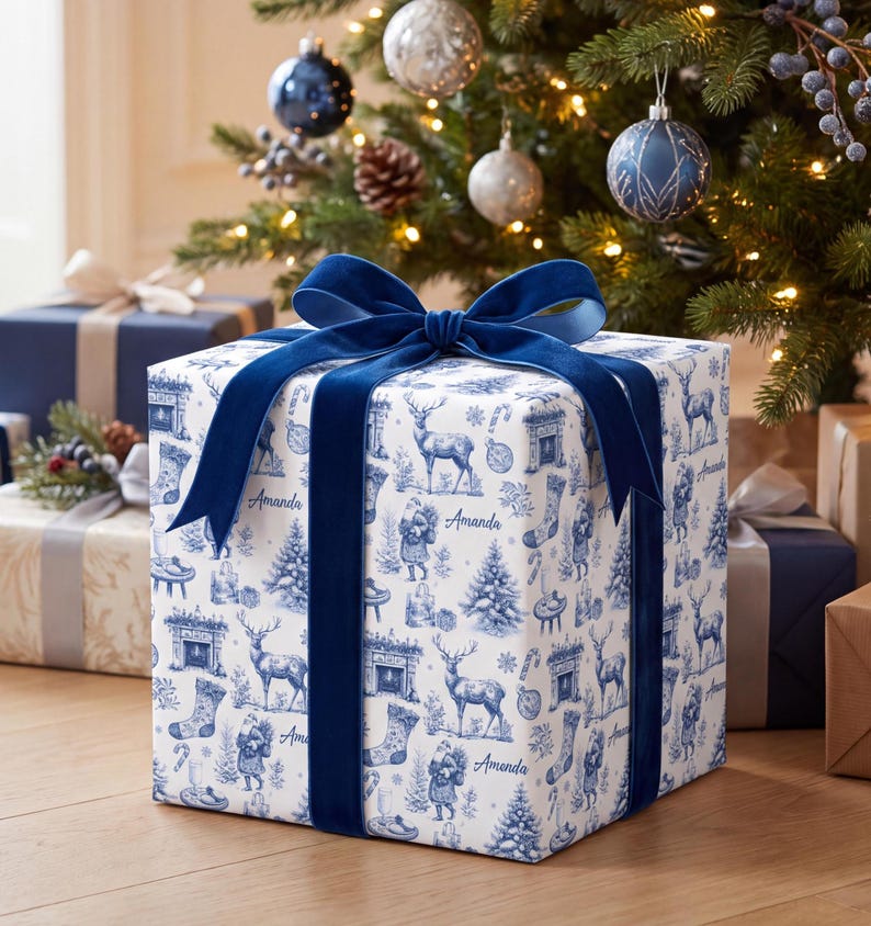 Puede incluir: Una caja de regalo cuadrada envuelta en papel blanco con un patr&oacute;n navide&ntilde;o azul estilo toile que muestra renos, Santa Claus, calcetines y chimeneas. El nombre Amanda est&aacute; impreso en el papel. Un lazo de terciopelo azul oscuro corona la caja.