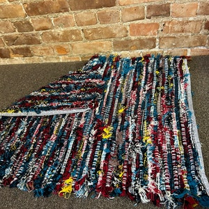 Könnte beinhalten: Ein bunter, handgewebter Teppich mit Fransen. Der Teppich besteht aus verschiedenen Blautönen, Rot, Weiß und Gelb.