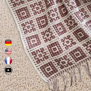 Puede incluir: Primer plano de una manta de ganchillo con un patrón geométrico en burdeos y crema. La manta tiene un borde con flecos y se muestra sobre una superficie texturizada de color claro. El patrón presenta cuadrados y rombos repetidos.