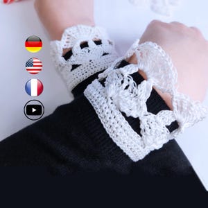 May include: White crochet wrist cuffs with a scalloped edge and floral design are worn over a black sleeve. The cuffs are handmade and add a delicate touch to any outfit.