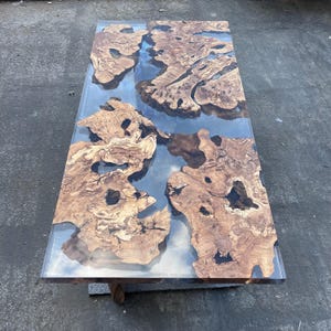 Puede incluir: Una mesa rectangular que presenta losas de madera natural con bordes irregulares, vetas intrincadas y nudos, incrustadas en una resina transparente y lisa que crea una apariencia de río sobre la superficie de la mesa.