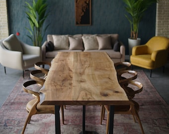 May include: A long, wooden table with a live edge sits in a modern living room. The table is surrounded by six chairs with brown leather seats and black metal legs. The table is made of natural wood with a light brown finish. The room is decorated with a neutral color palette and features a large window, a sofa, and two armchairs.