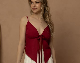 May include: A woman wearing a red top with a V-neck and tie detail, paired with wide-leg, off-white pants. The outfit is set against a neutral backdrop, with a wooden ladder and chair visible.