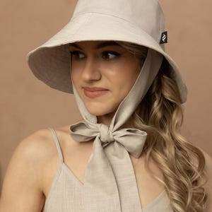 May include: A light beige sun hat with a wide brim and a matching fabric tie. The hat has a small black tag on the side. The woman is wearing a matching beige dress.