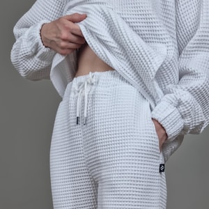 Pode incluir: Conjunto de roupa de descanso branco em malha waffle. O conjunto inclui uma parte superior de manga comprida e calças com cordão. O tecido tem um padrão texturizado em forma de grelha. As calças têm bolsos e uma pequena etiqueta com o logótipo.