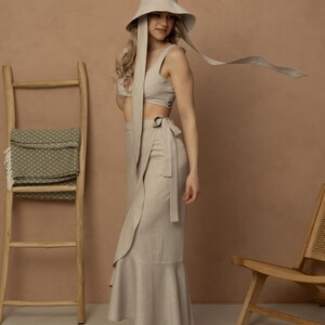 May include: A beige linen two-piece outfit, including a crop top and a wrap skirt with a ruffled hem. The outfit is paired with a matching wide-brimmed hat with long ties. The model is wearing black strappy heels. A wooden ladder and chair are in the background.