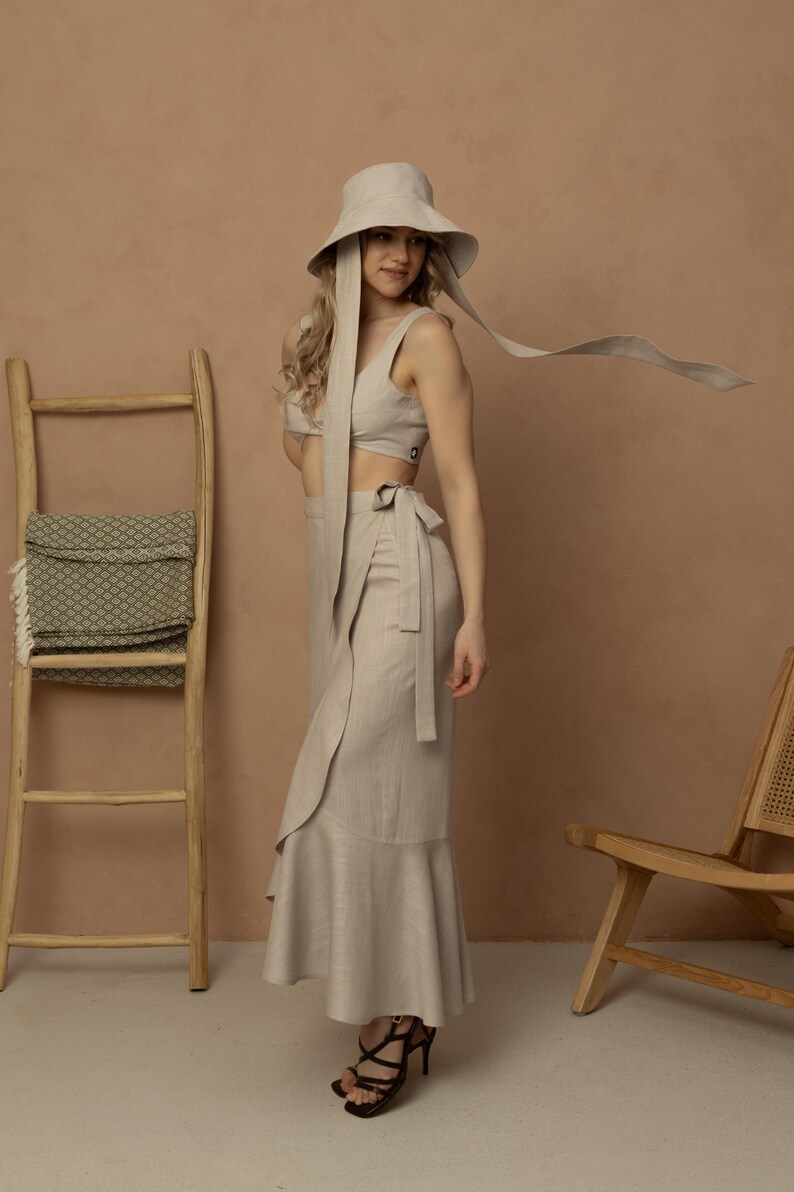 May include: A beige linen two-piece outfit, including a crop top and a wrap skirt with a ruffled hem. The outfit is paired with a matching wide-brimmed hat with long ties. The model is wearing black strappy heels. A wooden ladder and chair are in the background.