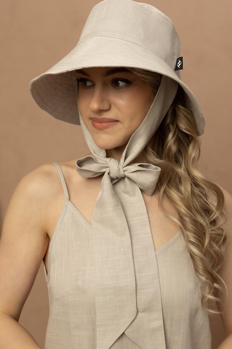 May include: A light beige sun hat with a wide brim and a matching fabric tie. The hat has a small black tag on the side. The woman is wearing a matching beige dress.