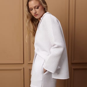 May include: A white waffle-weave jacket and shorts set. The jacket has a lapel collar and long sleeves. The shorts have pockets. The fabric has a textured, grid-like pattern. The outfit is a light, neutral color.