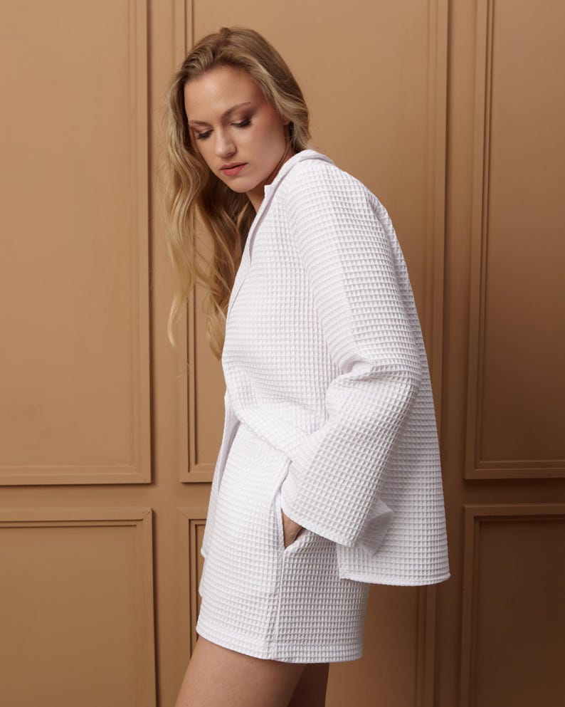 May include: A white waffle-weave jacket and shorts set. The jacket has a lapel collar and long sleeves. The shorts have pockets. The fabric has a textured, grid-like pattern. The outfit is a light, neutral color.