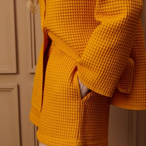 May include: Close-up of a mustard yellow waffle-weave robe with a matching belt. The robe has a pocket and is made of a textured fabric. The sleeves are wide and the robe is tied at the waist.