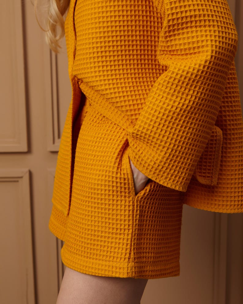 May include: Close-up of a mustard yellow waffle-weave robe with a matching belt. The robe has a pocket and is made of a textured fabric. The sleeves are wide and the robe is tied at the waist.