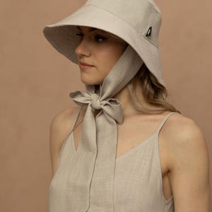 May include: A light beige sun hat with a wide brim and a matching fabric tie. The hat is made of a textured material and has a small black logo on the side. The tie is tied in a bow under the chin. The model is wearing a matching beige dress.