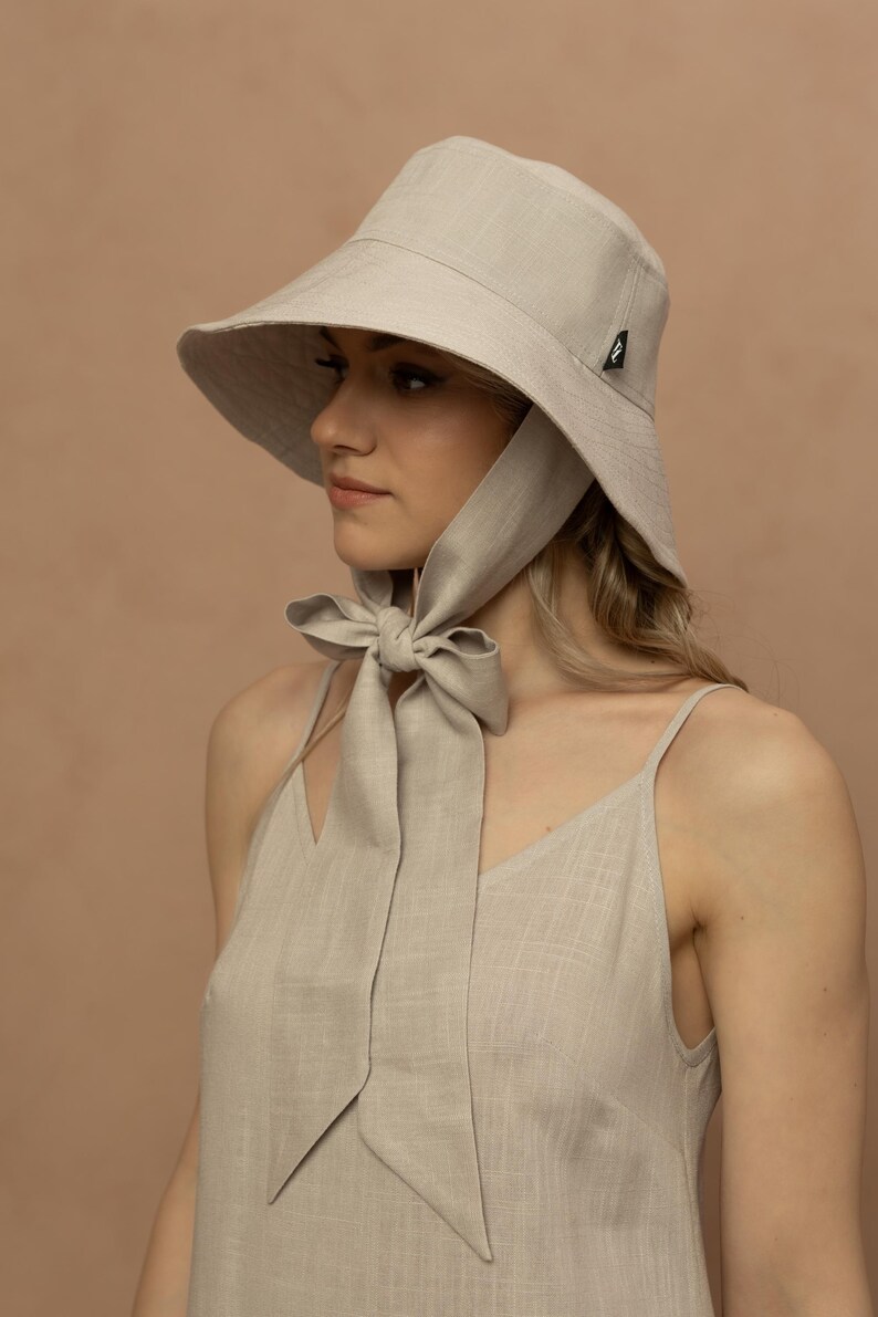 May include: A light beige sun hat with a wide brim and a matching fabric tie. The hat is made of a textured material and has a small black logo on the side. The tie is tied in a bow under the chin. The model is wearing a matching beige dress.