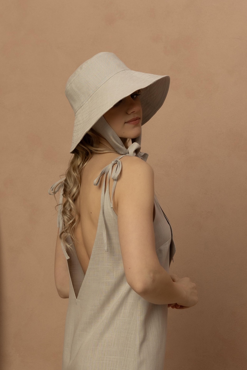 May include: A light beige sun hat with a wide brim and a matching dress. The dress has a low back and tie straps. The hat has a chin strap. The outfit is made of a linen-like fabric.