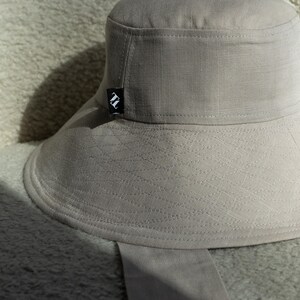 May include: A light gray bucket hat with a wide brim and a small black and white logo on the side. The hat is made of a textured fabric and has a matching fabric strap.