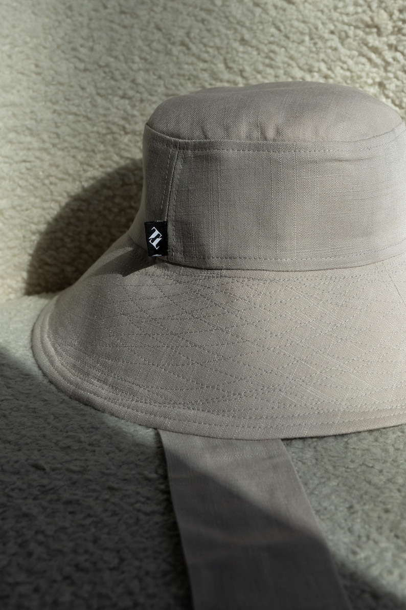 May include: A light gray bucket hat with a wide brim and a small black and white logo on the side. The hat is made of a textured fabric and has a matching fabric strap.