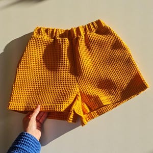 May include: Mustard yellow waffle-weave shorts. The shorts have an elastic waistband and a textured surface. The garment is displayed on a light-colored surface, with a hand wearing a blue sleeve holding the shorts.