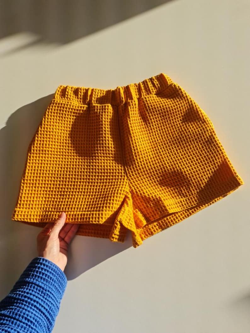 May include: Mustard yellow waffle-weave shorts. The shorts have an elastic waistband and a textured surface. The garment is displayed on a light-colored surface, with a hand wearing a blue sleeve holding the shorts.