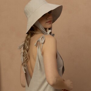 May include: A light beige sun hat with a wide brim and a matching dress. The dress has a low back and tie straps. The hat has a chin strap. The outfit is made of a linen-like fabric.